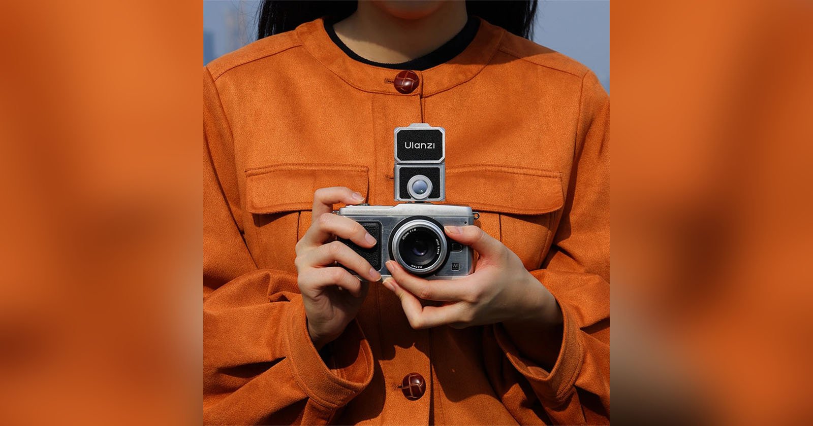 A person wearing an orange coat is holding a camera with both hands. The camera has a Ulanzi-branded flash or accessory mounted on top. The person's face is partially out of the frame.