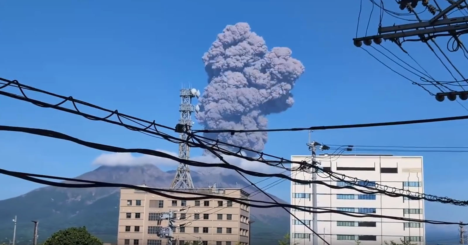 A volcano erupts, sending a large cloud of ash into the clear blue sky. The eruption is visible behind buildings and numerous power lines in the foreground. The scene is framed by urban structures, highlighting the contrast between nature and civilization.
