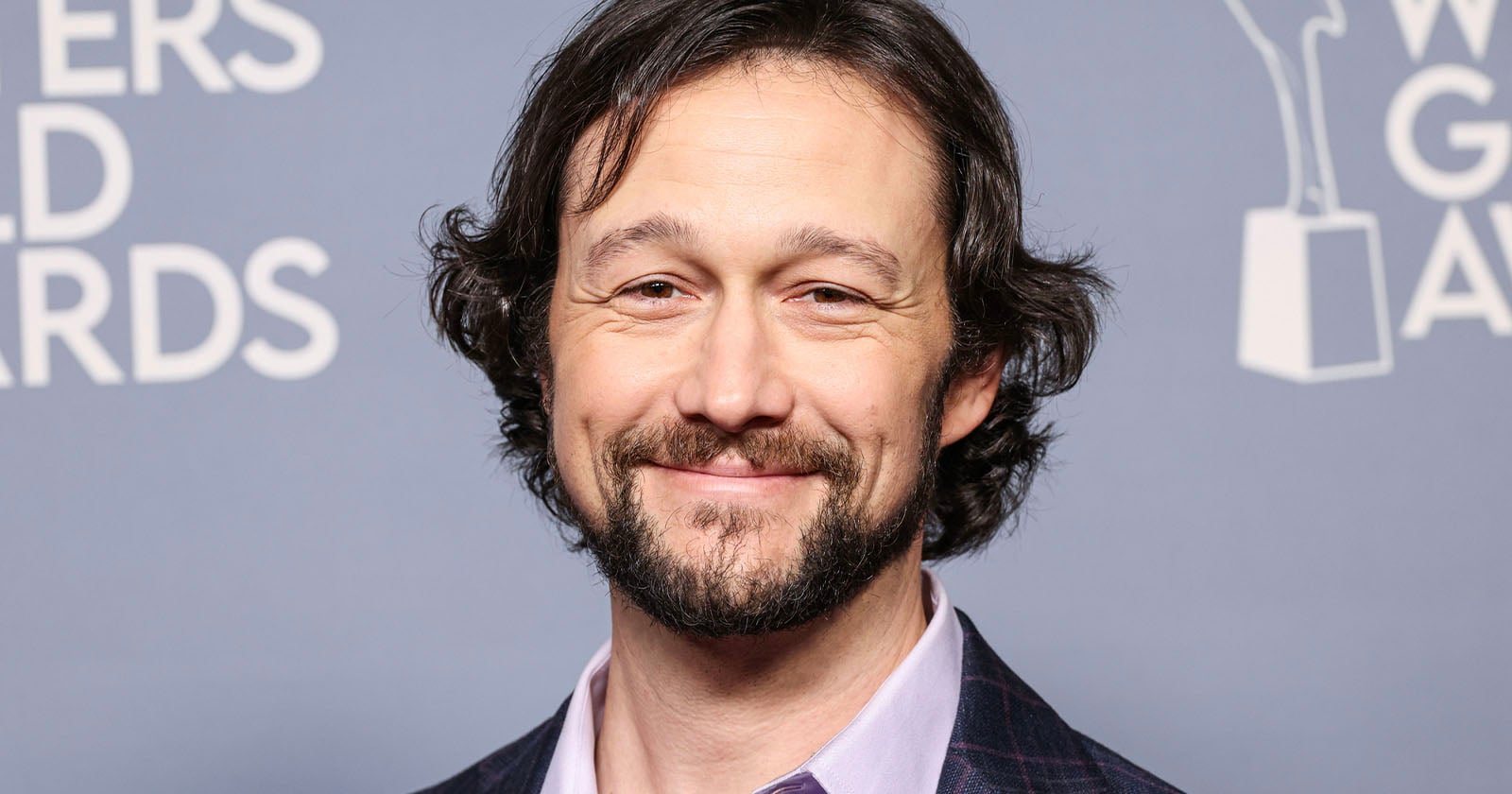 A man with wavy dark hair and a beard smiles in front of a gray backdrop with white text and an award statue graphic. He is wearing a light pink collared shirt and a plaid jacket.