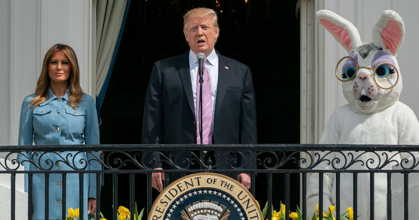 Three figures stand on a balcony: a woman in a blue dress, a man in a suit speaking at a podium with the presidential seal, and a person in a large white rabbit costume with glasses. Yellow flowers line the balcony railing.