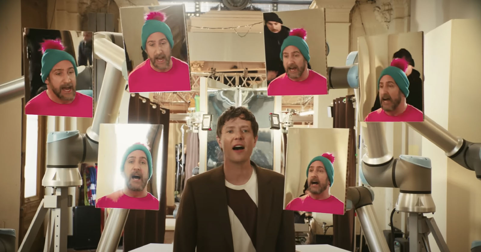 A person sings in the center while surrounded by six screens showing a man in a bright pink shirt and blue winter hat. Robotic arms hold the screens in a brightly lit room with curtains and lights in the background.