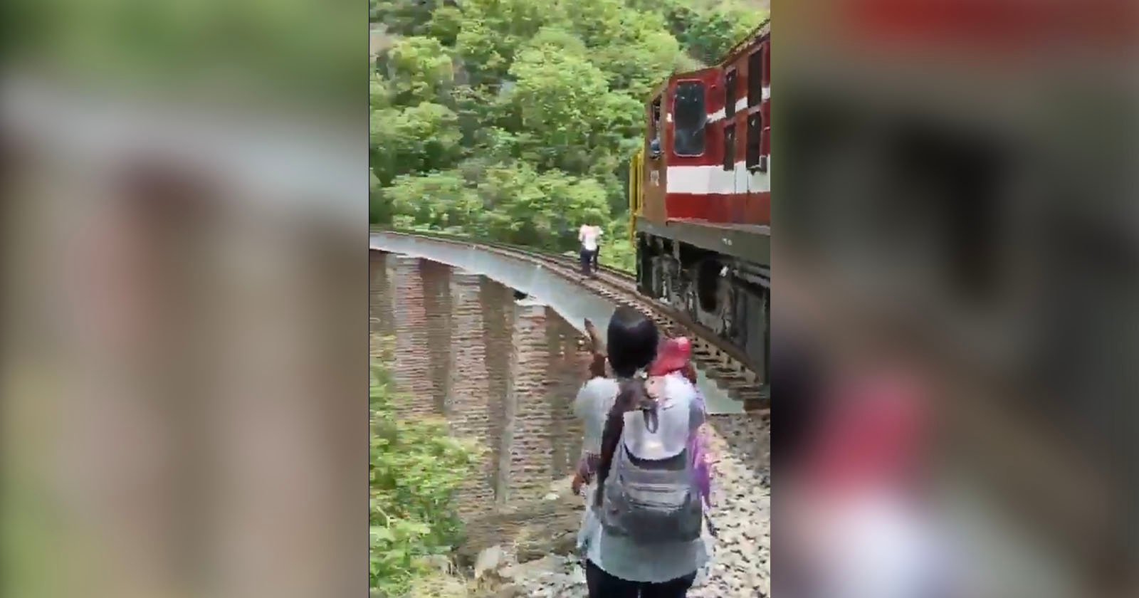 Two people carrying backpacks walk along a narrow train track beside a moving red and white train. The track is elevated with a steep drop on one side, surrounded by dense green foliage. Another person is walking ahead of them on the tracks.