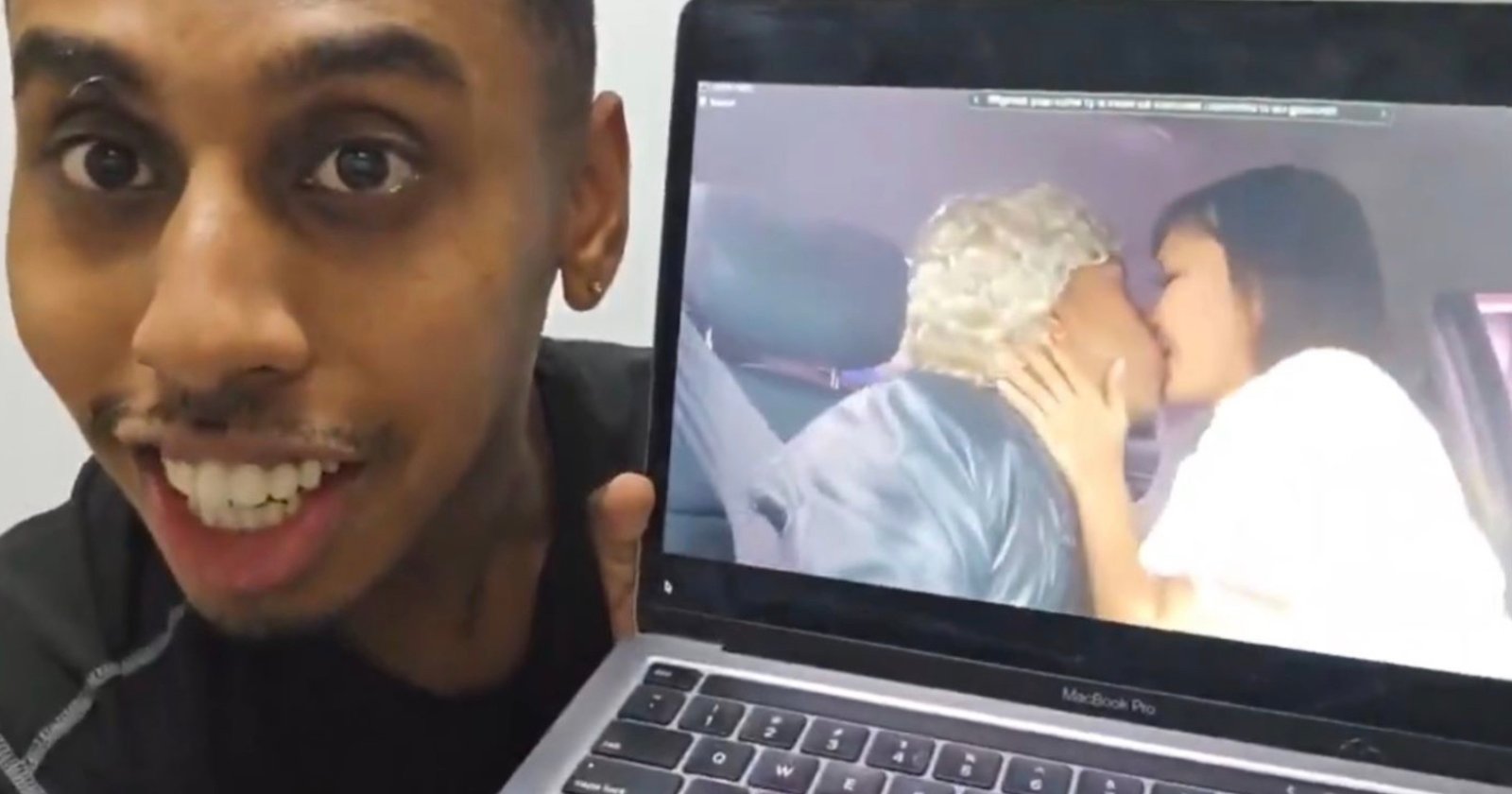 A person smiling and holding a laptop displaying a video. In the video, two people are seen kissing; one has blonde hair and the other has long brown hair.
