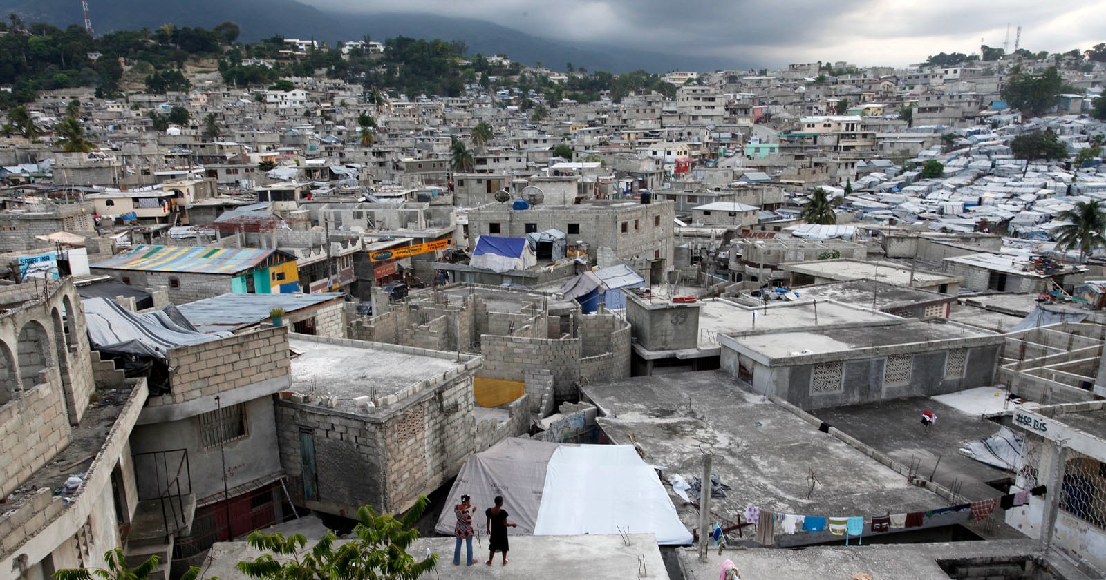 A densely packed hillside cityscape with concrete buildings and makeshift structures covered in tarps, showing signs of crowding and poverty under a cloudy sky, with mountains visible in the background.
