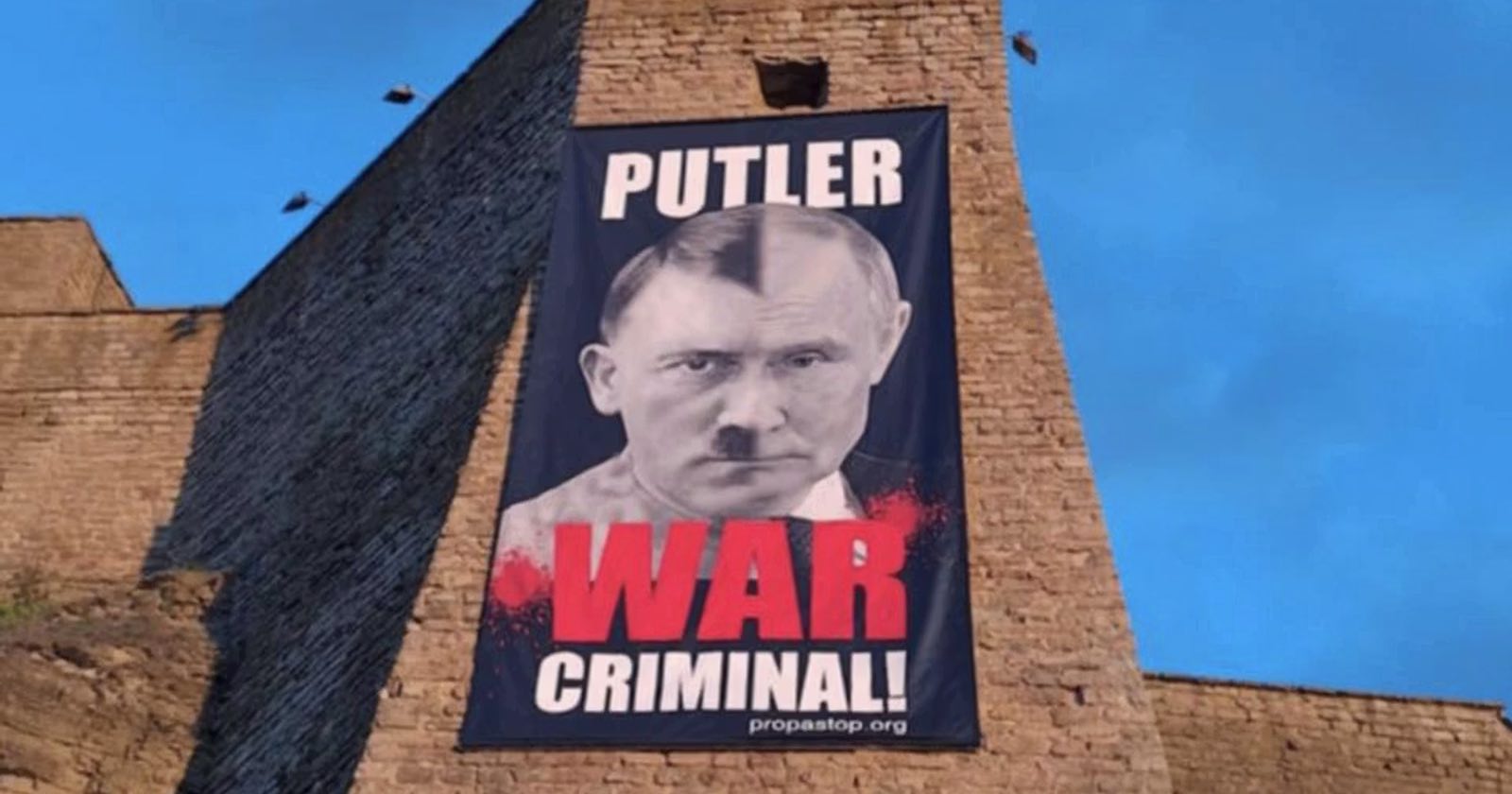 A large banner hangs on a stone wall, featuring a photoshopped image combining Vladimir Putin’s and Adolf Hitler’s faces, with the words PUTLER WAR CRIMINAL! in bold letters below.