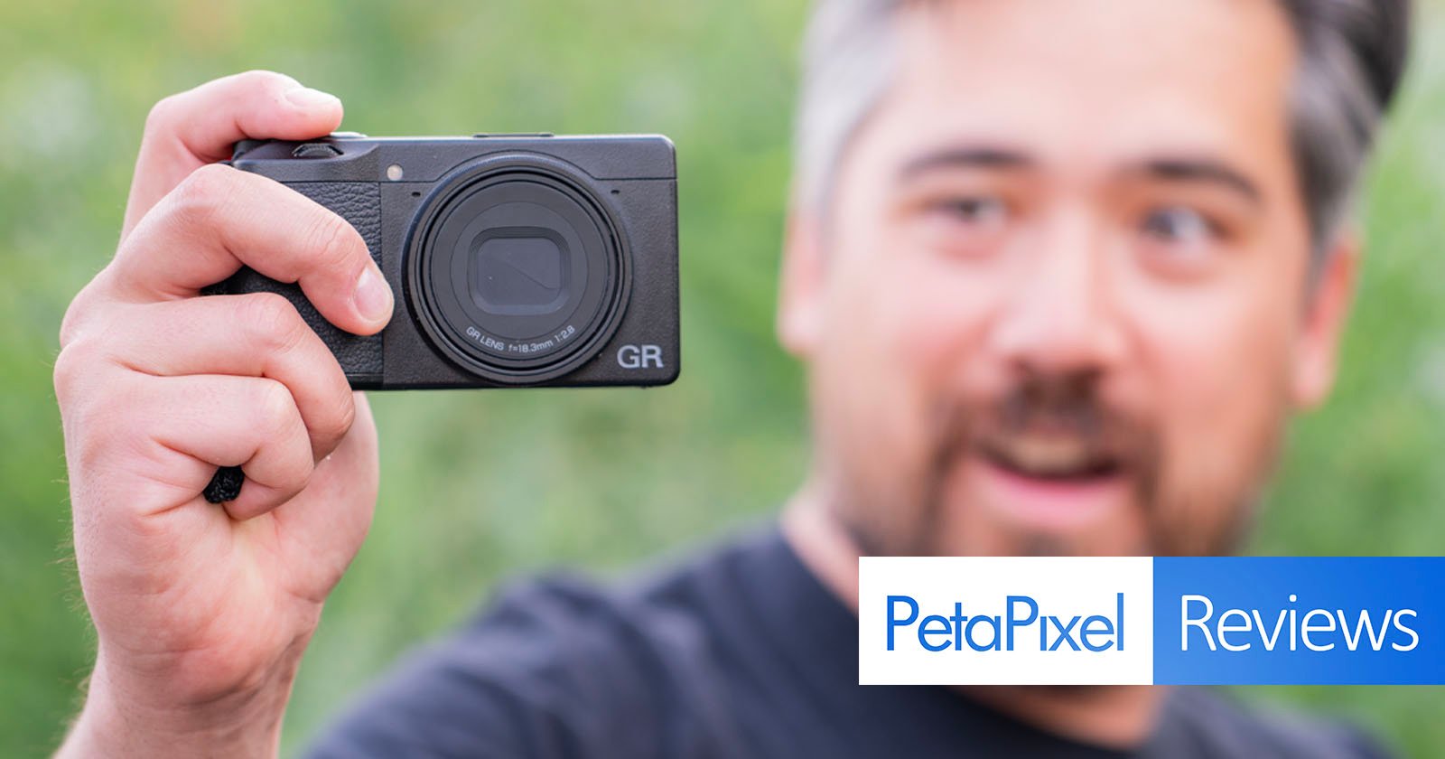 A person holds up a black Ricoh GR camera in their left hand, displaying it prominently. The background is blurred green foliage. In the bottom right corner, there's a banner with the text PetaPixel Reviews. The person is smiling and partially out of focus.