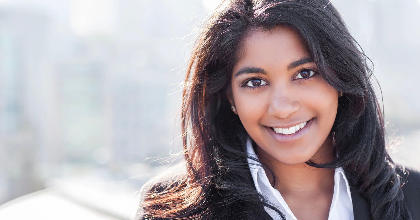 Smiling person with long dark hair stands outdoors, wearing a white shirt and dark jacket. The background is blurred, suggesting an urban setting. They are looking directly at the camera, with a bright, cheerful expression.