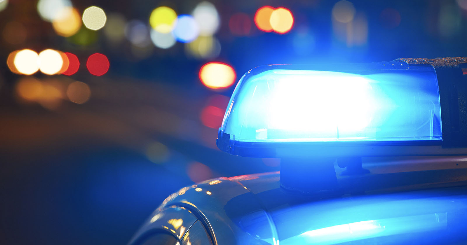 Close-up of a blue police car siren flashing brightly at night, with blurred colorful lights in the background, suggesting a busy urban street scene.