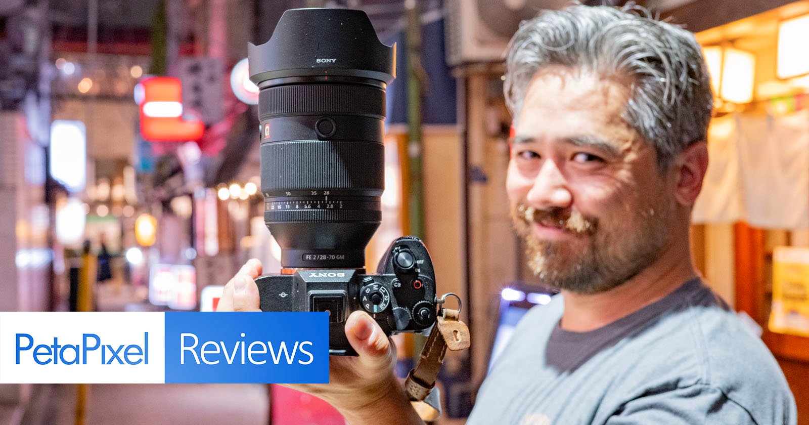 A person holding a Sony camera with a large lens in a dimly lit street, smiling slightly. The background features illuminated signs and blurred lights. Overlay text reads PetaPixel Reviews.