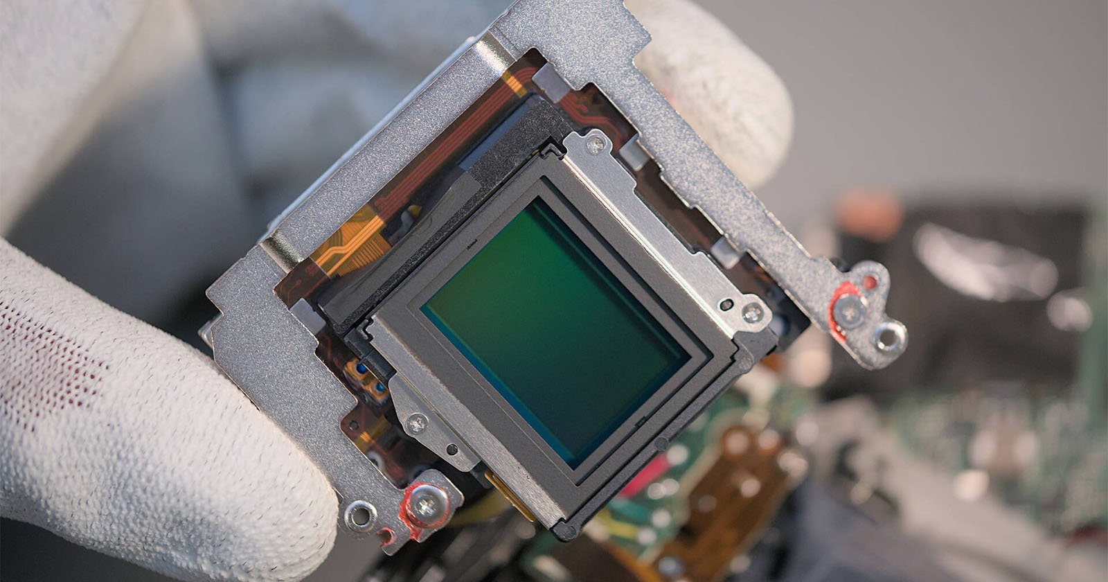 A gloved hand holds a digital camera image sensor surrounded by a metallic frame, showing intricate electronic components and circuitry.