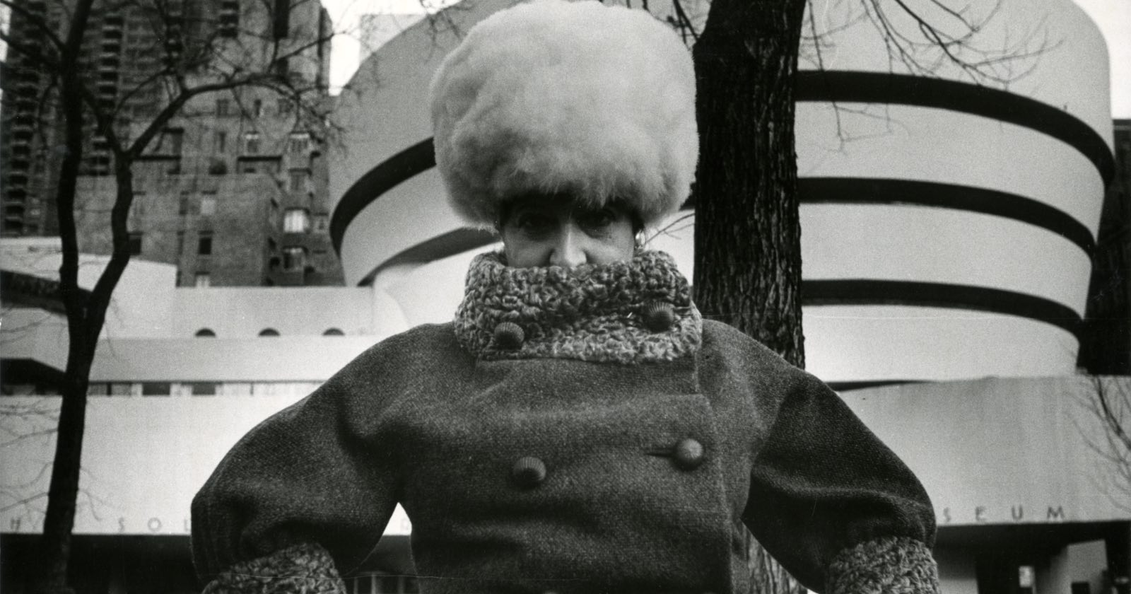 A person in a thick, high-collared coat and large, fluffy hat stands outside in winter, with the circular architecture of the Guggenheim Museum and bare trees in the background.