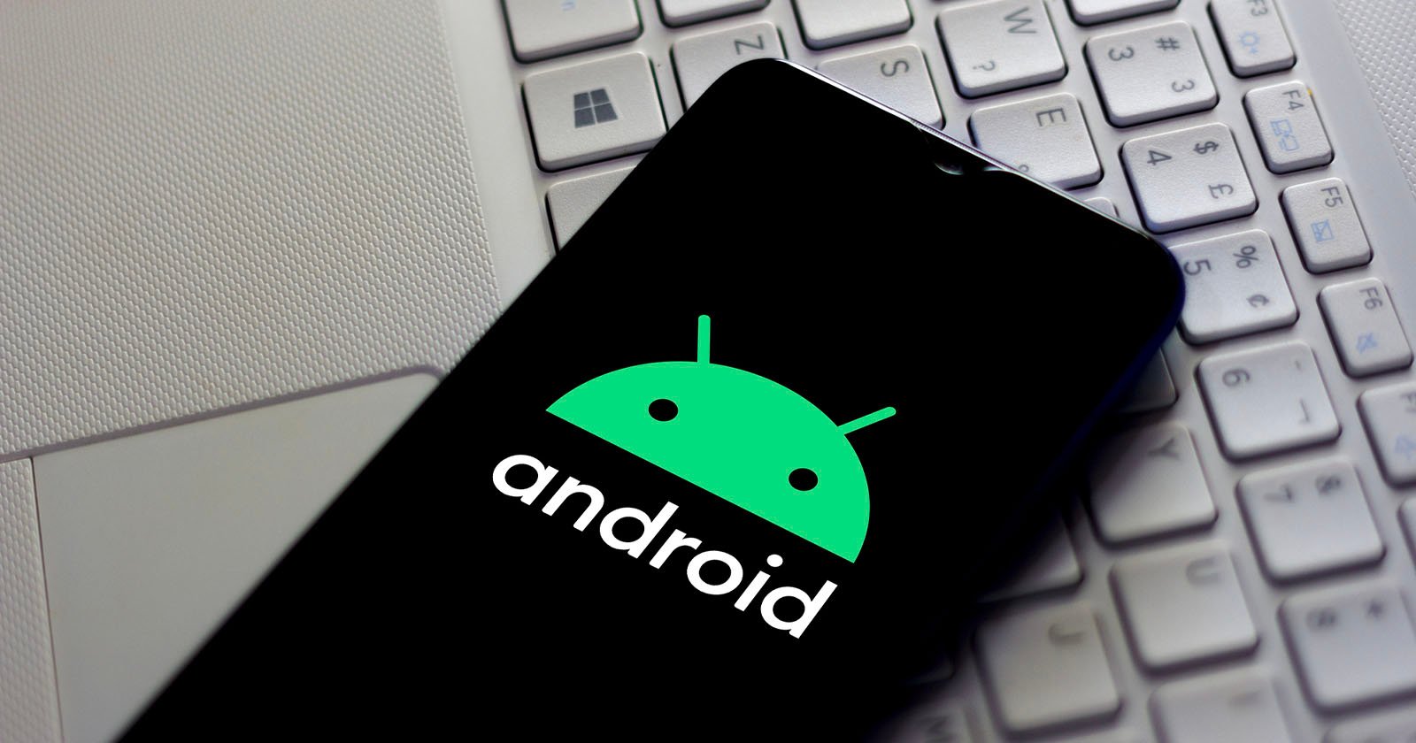 A smartphone displays the green Android logo and the word android on its screen, resting on a silver laptop keyboard.