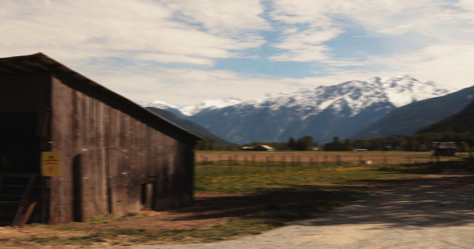 A rustic wooden barn stands on a grassy field with a backdrop of snow-capped mountains under a partly cloudy sky. A dirt path runs alongside the barn.