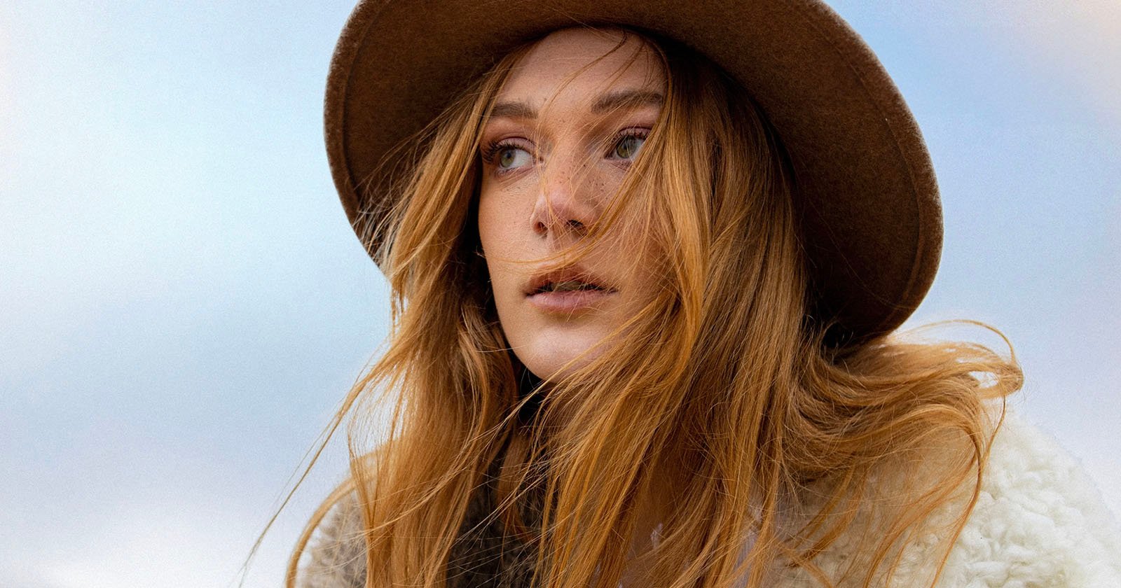 Woman with long, flowing red hair wearing a brown hat and cream coat gazes thoughtfully into the distance against a blue sky background.