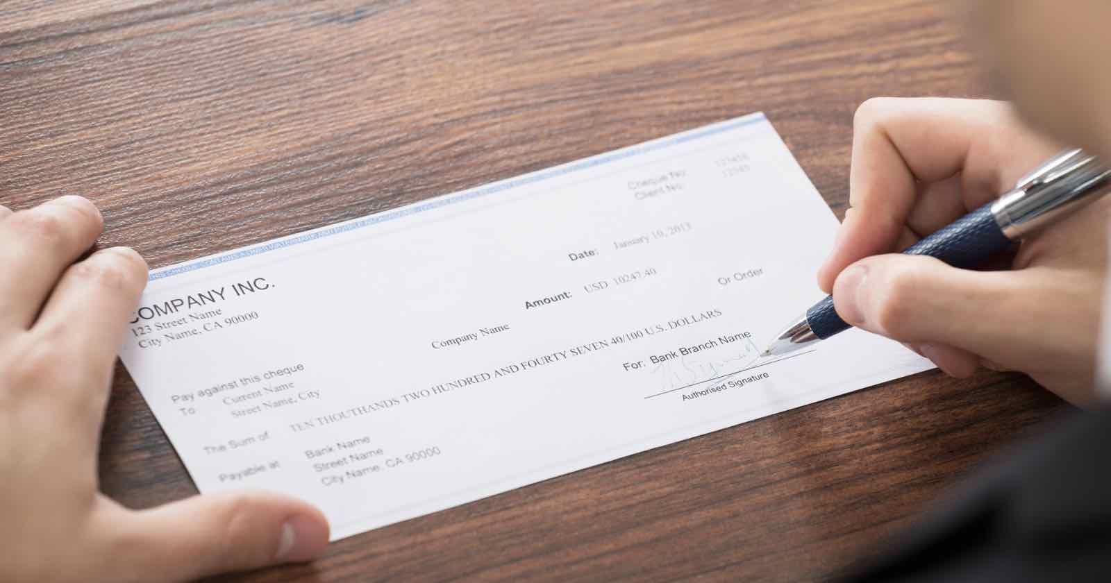 A person signs a check on a wooden table. The check, issued by Company Inc., is addressed to an individual, with a date of January 15, 2023. The amount is two hundred forty-seven dollars. A pen is held in the person's right hand.