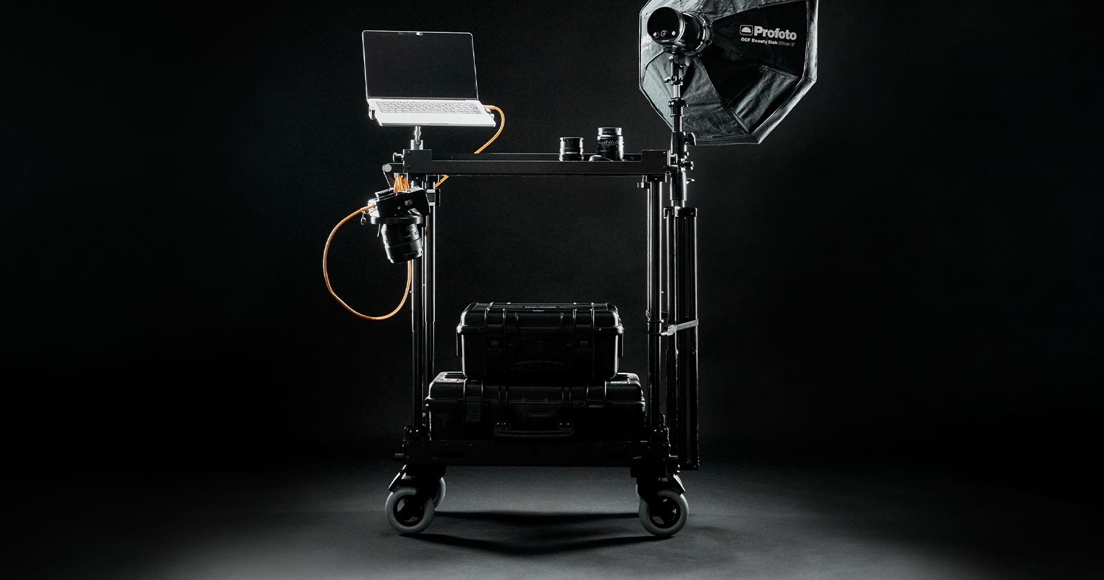 A mobile photography setup on a black background, featuring a laptop on a stand, a camera with an orange cable, a large black softbox light, several lenses, and a protective black carrying case on a wheeled cart.