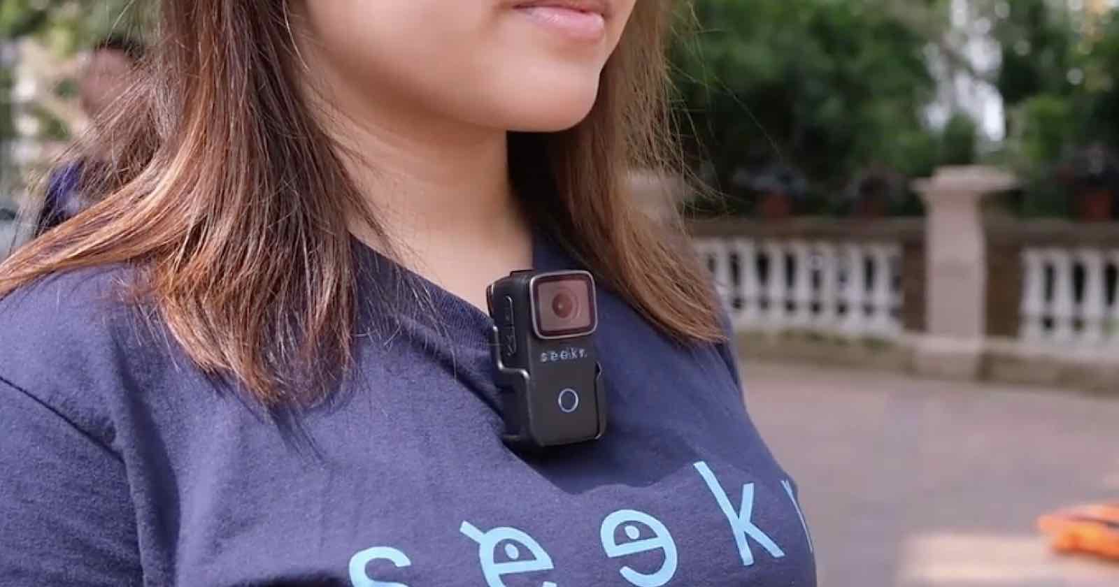 A person wearing a navy blue T-shirt with the word seek printed on it is standing outside. A small black camera is clipped to the front of the T-shirt. Trees and a fence are visible in the background.