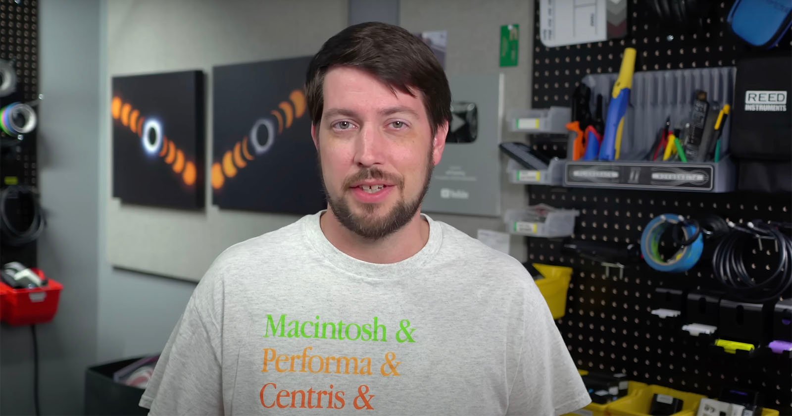 A person with short dark hair and a beard stands in a workshop-like setting. They are wearing a light gray T-shirt with green and orange text that reads Macintosh & Performa & Centris & Quadra. Various tools, cables, and supplies are organized on shelves and pegboards behind them.