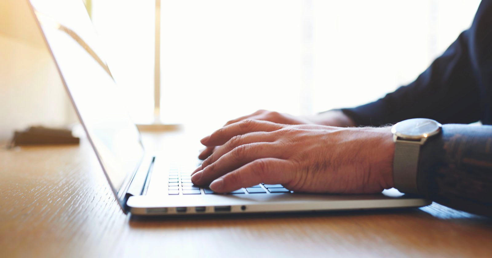 Close-up of a person typing on a laptop. The person's hands are positioned on the keyboard, with a wristwatch visible on the left wrist. The background is bright, suggesting daylight, with light streaming in through a window. The surface looks like a wooden desk.