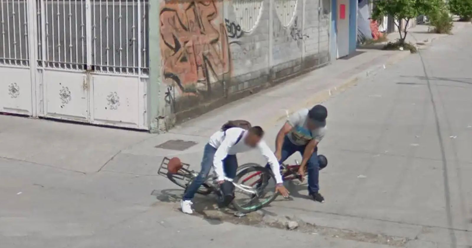 Two people are picking up their bicycles after falling into a pothole on a city street. One wears a white shirt and backpack; the other wears a hat and gray shirt. A gated building and graffiti-covered wall are visible in the background.
