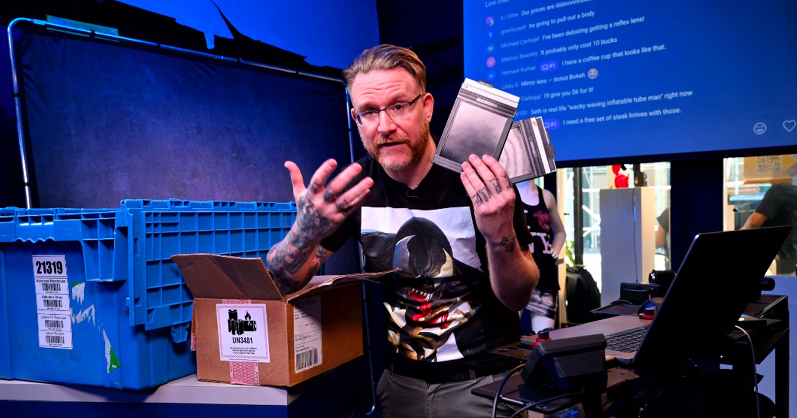 A person with glasses and tattoos holds metal plates while sitting at a desk with a laptop, blue storage bins, and a cardboard box in a brightly lit, tech-filled room. A chat screen is visible in the background.