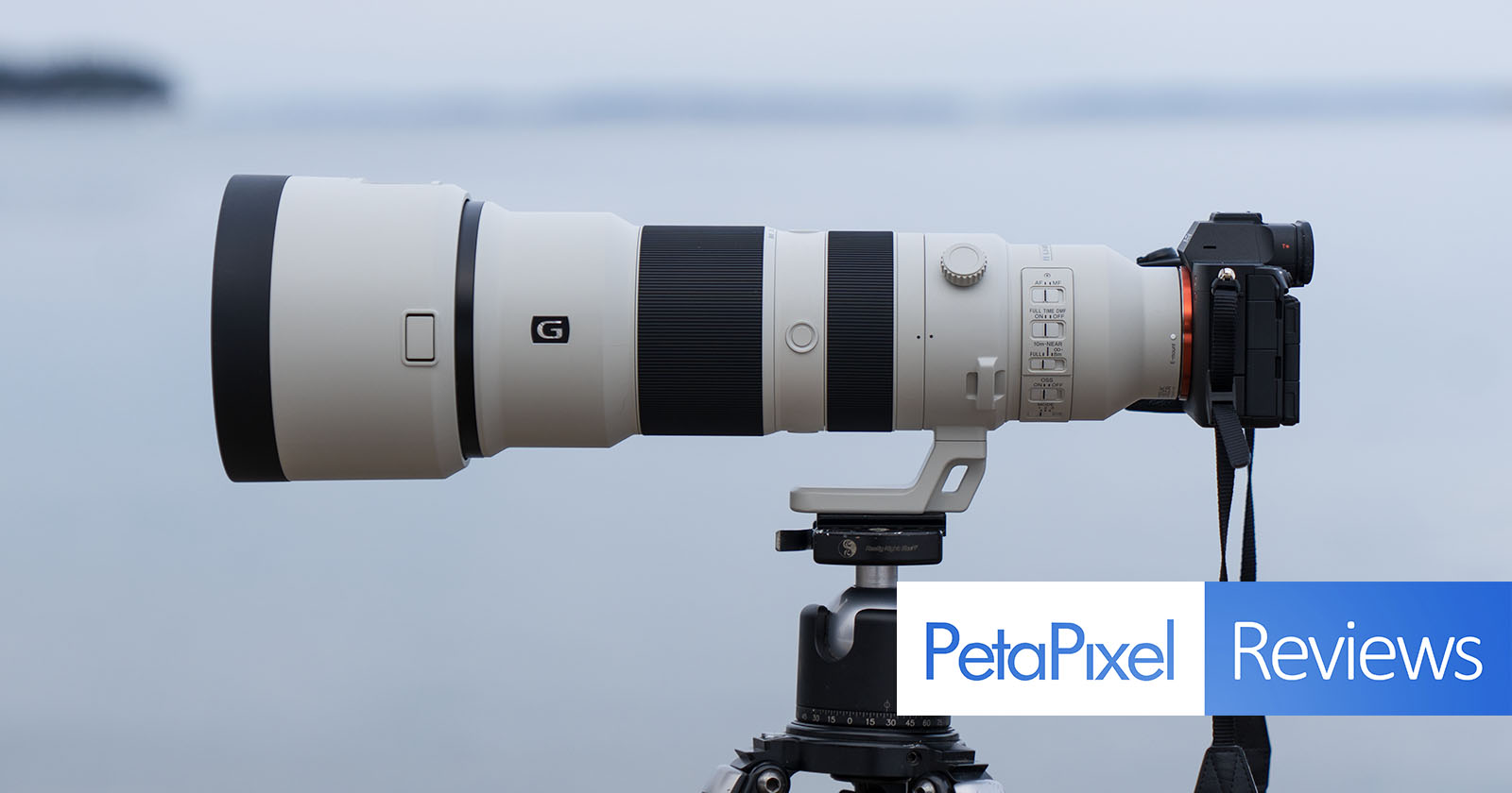 A large telephoto camera lens mounted on a tripod is shown. The lens is attached to a camera, and the setup is positioned to capture distant images. The word PetaPixel appears in blue alongside the word Reviews in white against a blue background.