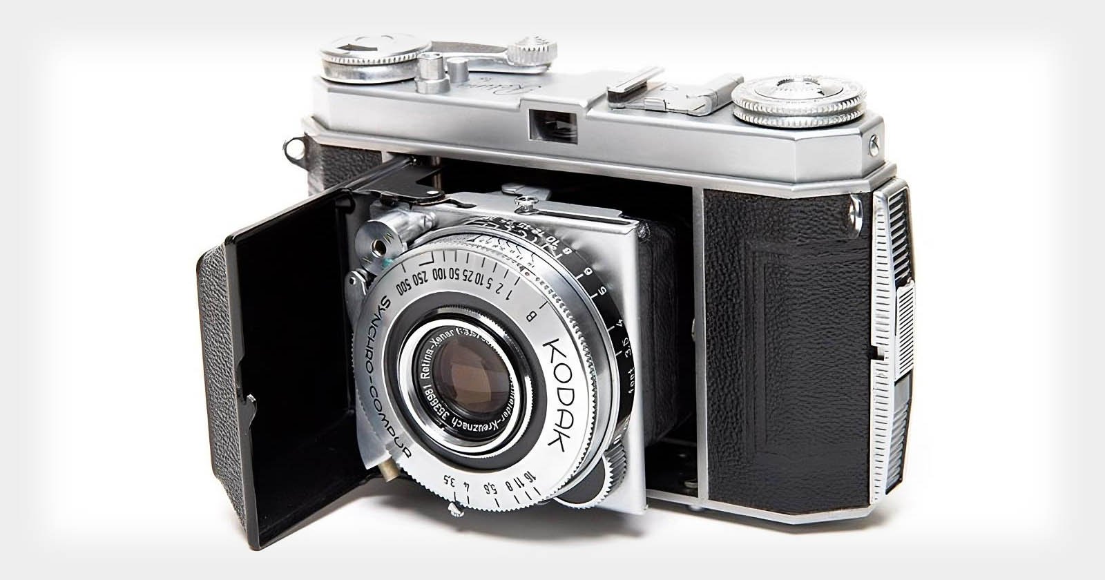 A vintage Kodak Retina IIc camera with a folding bellows design. The lens is extended, showing detailed dials and markings. The body is metal with black textured panels.