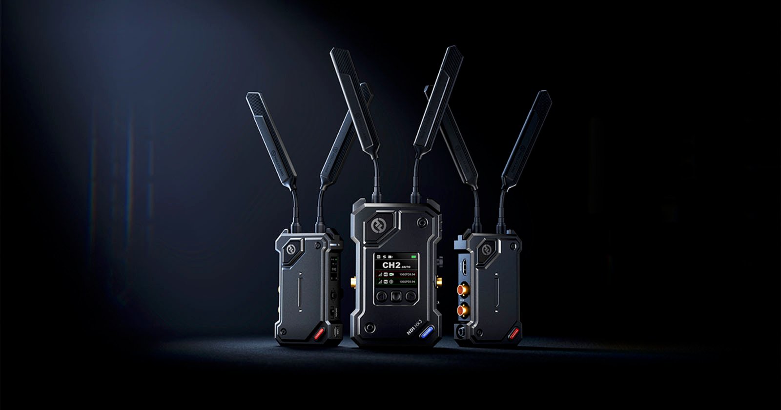 Three black wireless transmitter units with multiple antennas are standing on a dark surface against a black background. The central unit displays a digital screen with channel information.