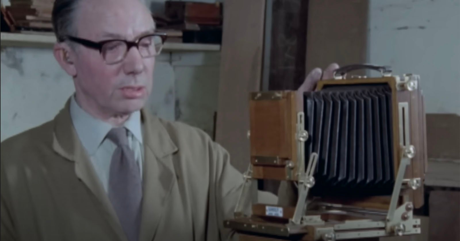 A man wearing glasses and a beige coat stands beside a large, vintage wooden camera with a bellows, inside a workshop or studio with shelves in the background.