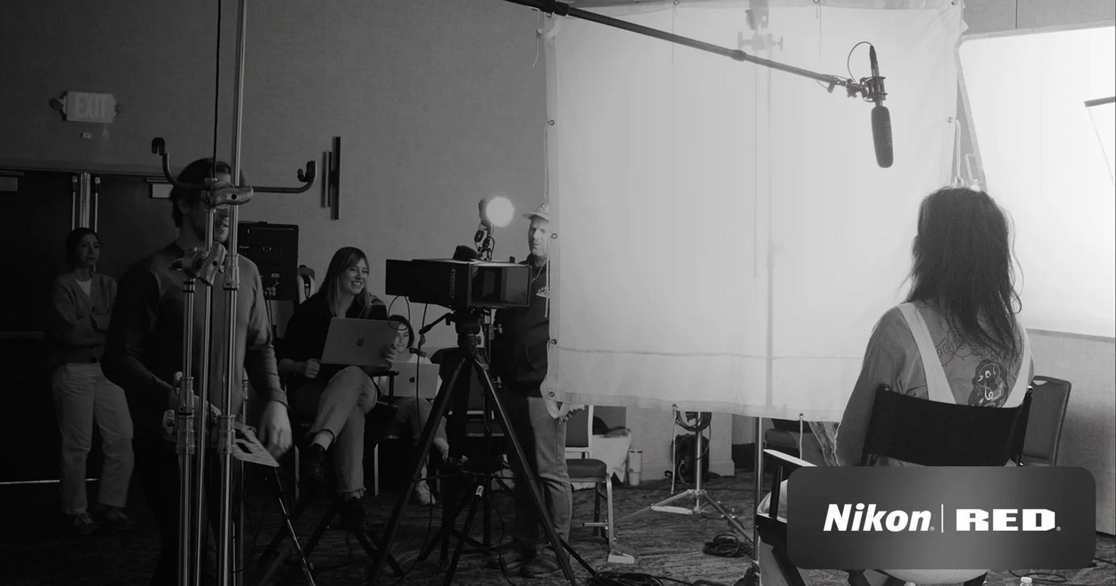 A black and white image of a video recording setup in a room. A person, sitting in a chair, is lit by studio lights facing them. Camera equipment and crew are visible, including a person holding a clipboard in the background. Nikon RED logo is in the corner.