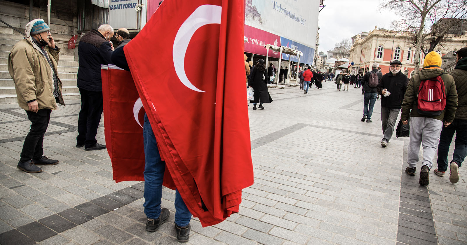 A person stands in a plaza holding several large Turkish flags, obscuring their body. People walk past on the gray cobblestone street, some wearing winter clothing. A building with columns is in the background. The scene feels casual and busy.