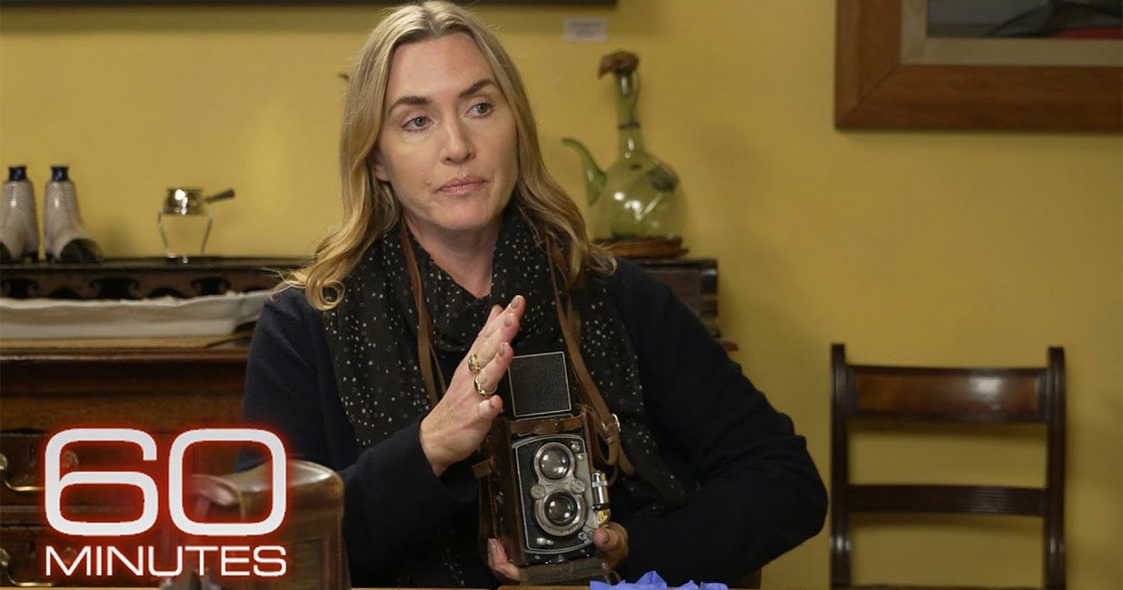 A woman with long blonde hair sits indoors, holding a vintage camera. She is surrounded by various objects and appears to be speaking. The 60 Minutes logo is visible in the bottom left corner.