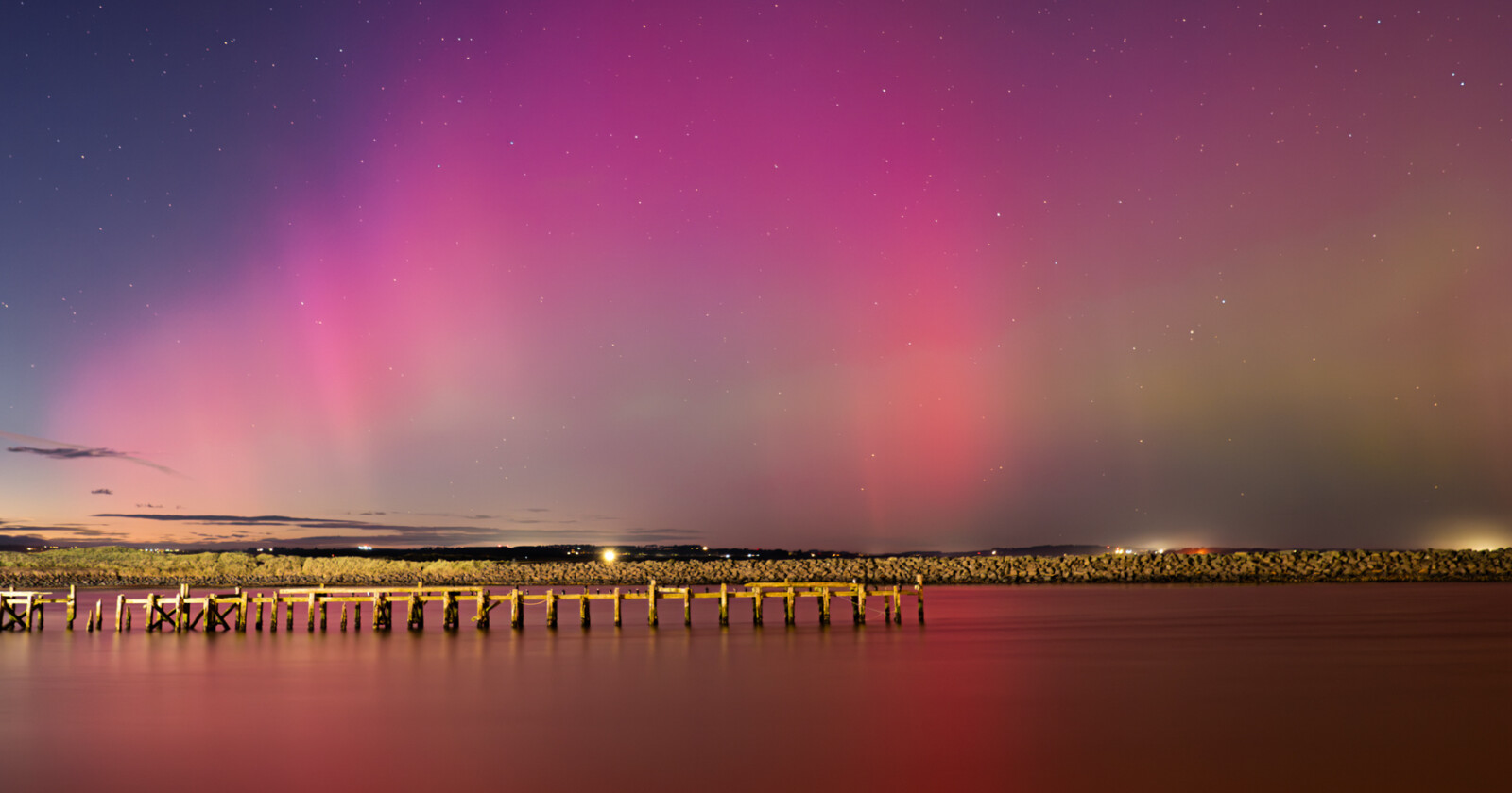 A vivid aurora with pink and purple lights illuminates the night sky above calm water and a wooden pier, with a rocky breakwater in the background.