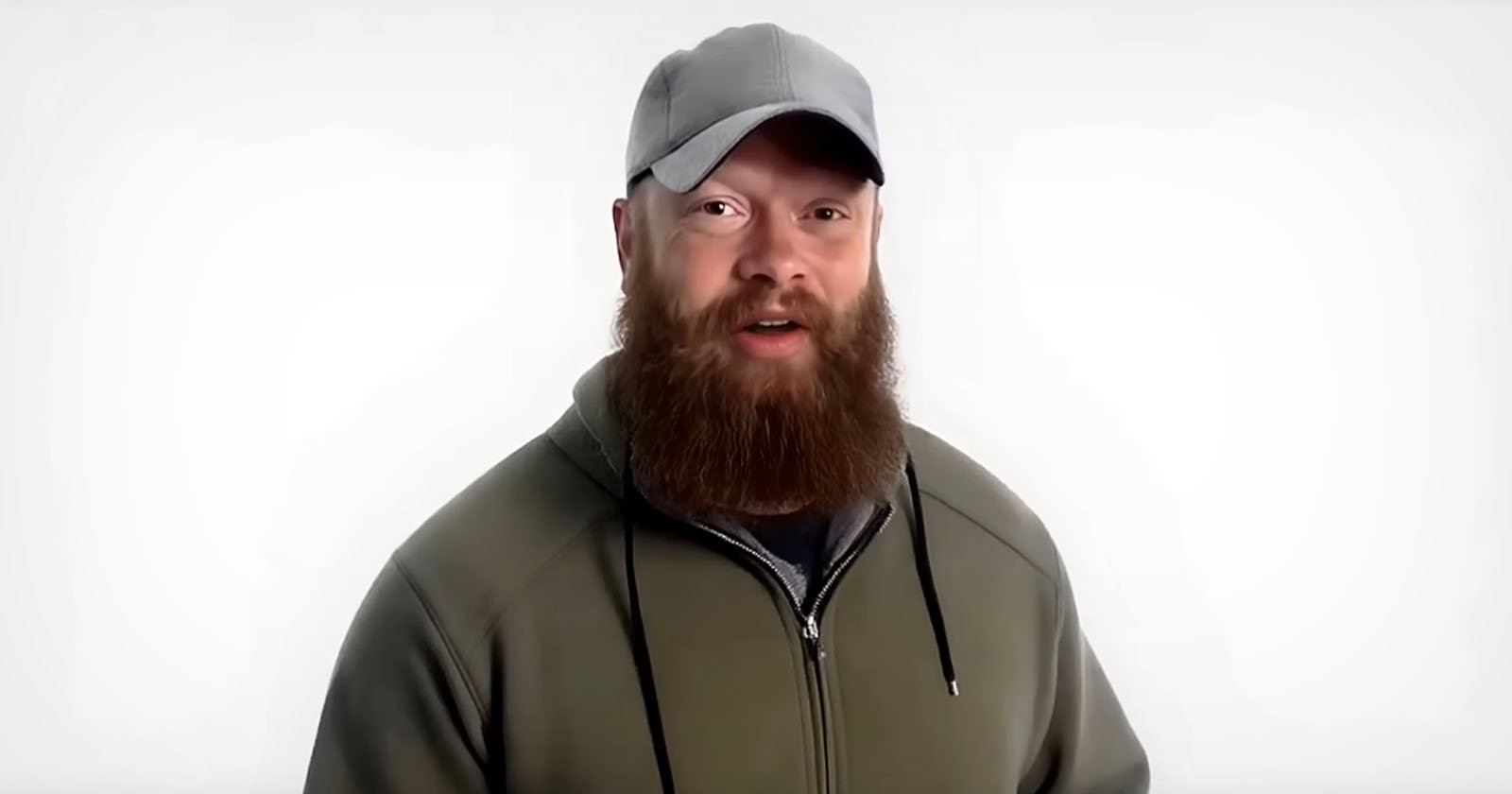 A man with a thick brown beard and mustache wears a gray baseball cap and an olive green hoodie. He stands in front of a plain white background, looking toward the camera.