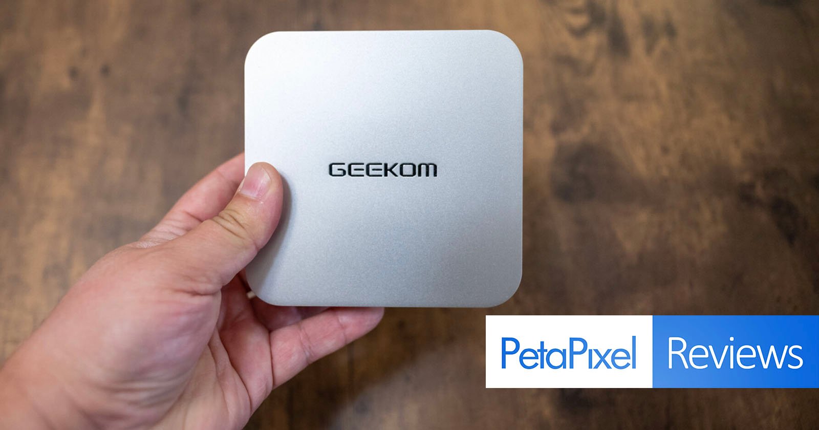 Hand holding a compact silver device with the brand name GEEKOM on top. The background is a wooden surface, and the PetaPixel Reviews logo is visible at the bottom right.