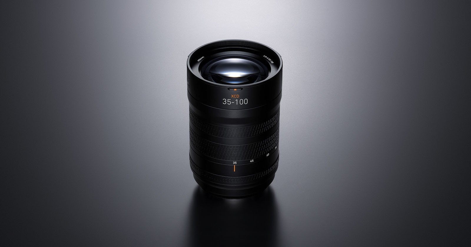 A black camera lens with “35–100” marking is standing upright on a smooth, dark surface under soft lighting, highlighting its textured grip and glass elements.