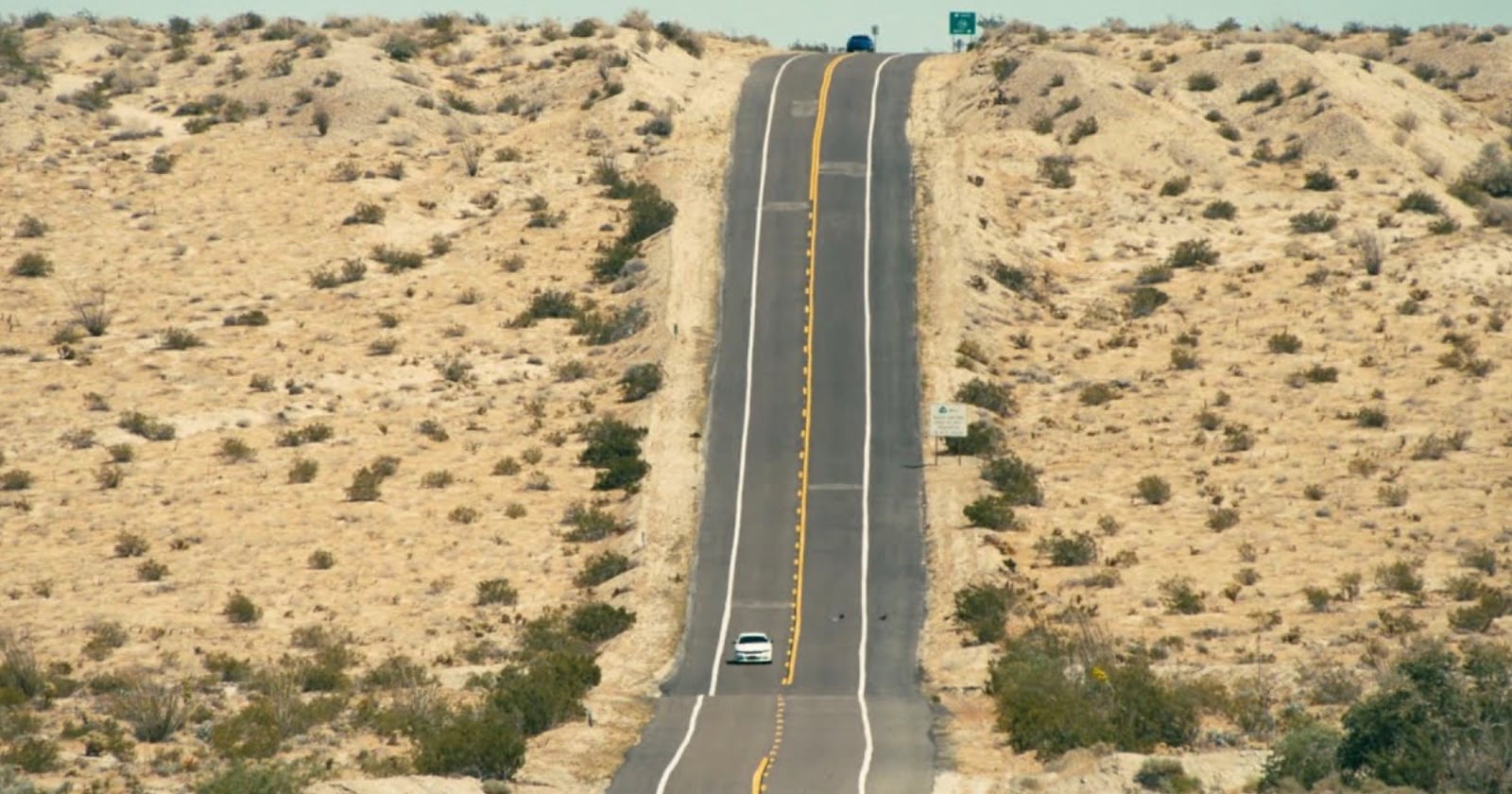 A long, straight road stretches through a dry, sandy desert landscape with sparse shrubs. One white car drives towards the horizon, while another vehicle is barely visible at the top of a hill ahead.