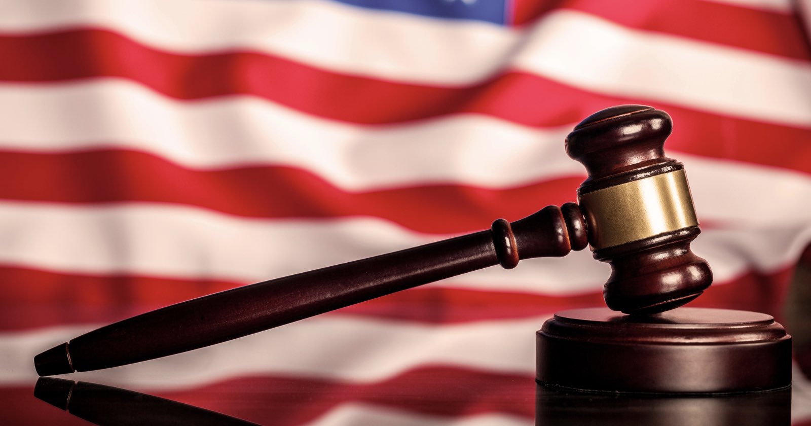 A wooden judge’s gavel rests on a sound block with a blurred American flag in the background.