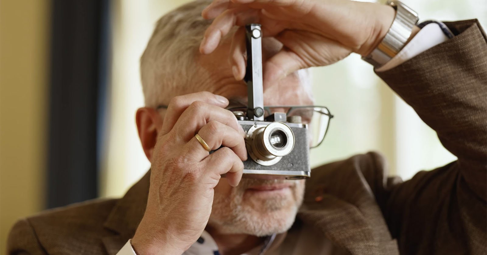 An older man with gray hair and glasses holds a vintage camera up to his face, preparing to take a photo. He wears a brown jacket, a gold ring, and a silver watch. The background is softly blurred.