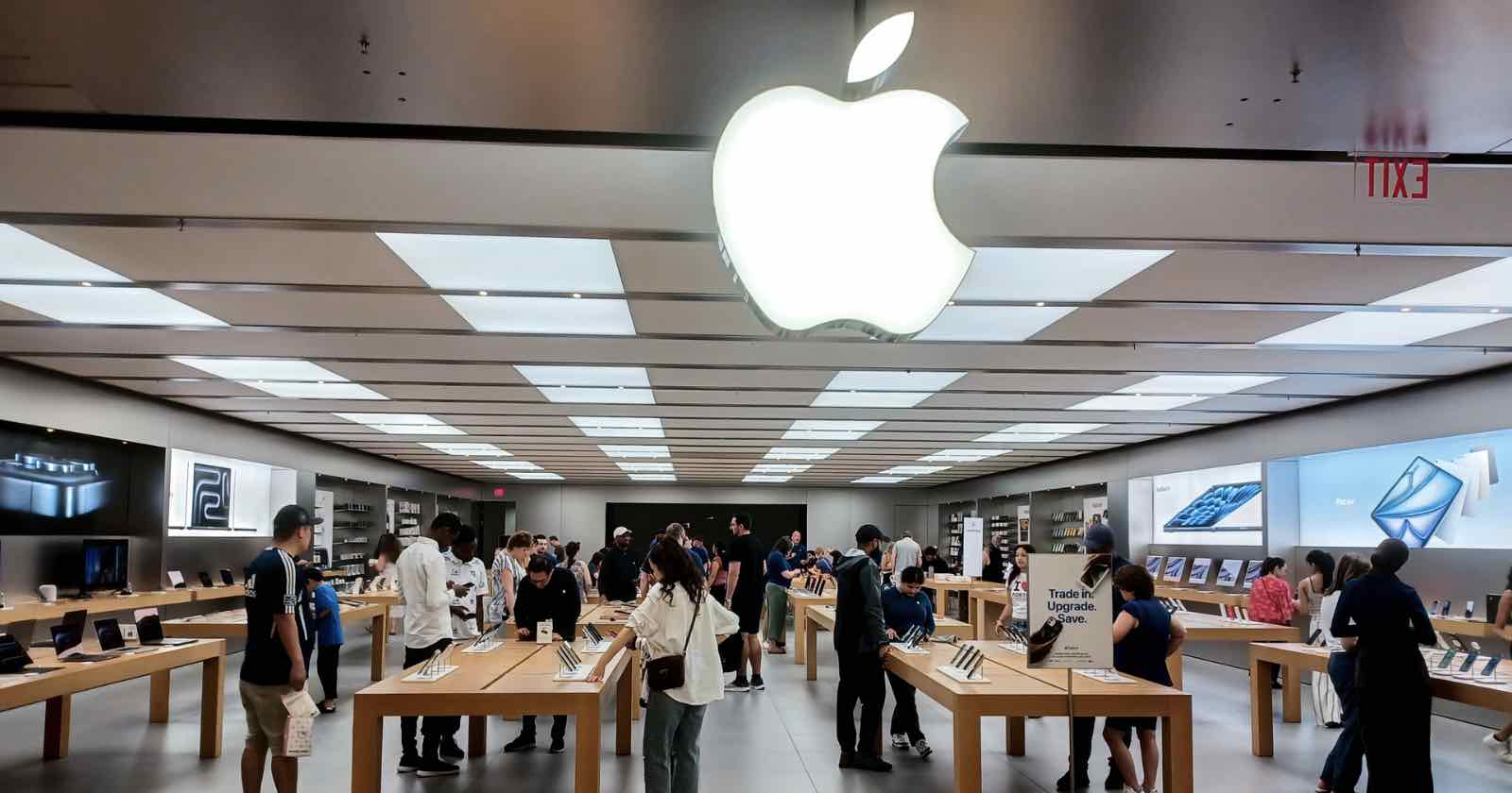 Inside an Apple Store, people browse products displayed on wooden tables. Large Apple logo is illuminated on the ceiling. Shelves filled with boxes and screens line the walls, and a busy atmosphere prevails with shoppers and staff interacting.