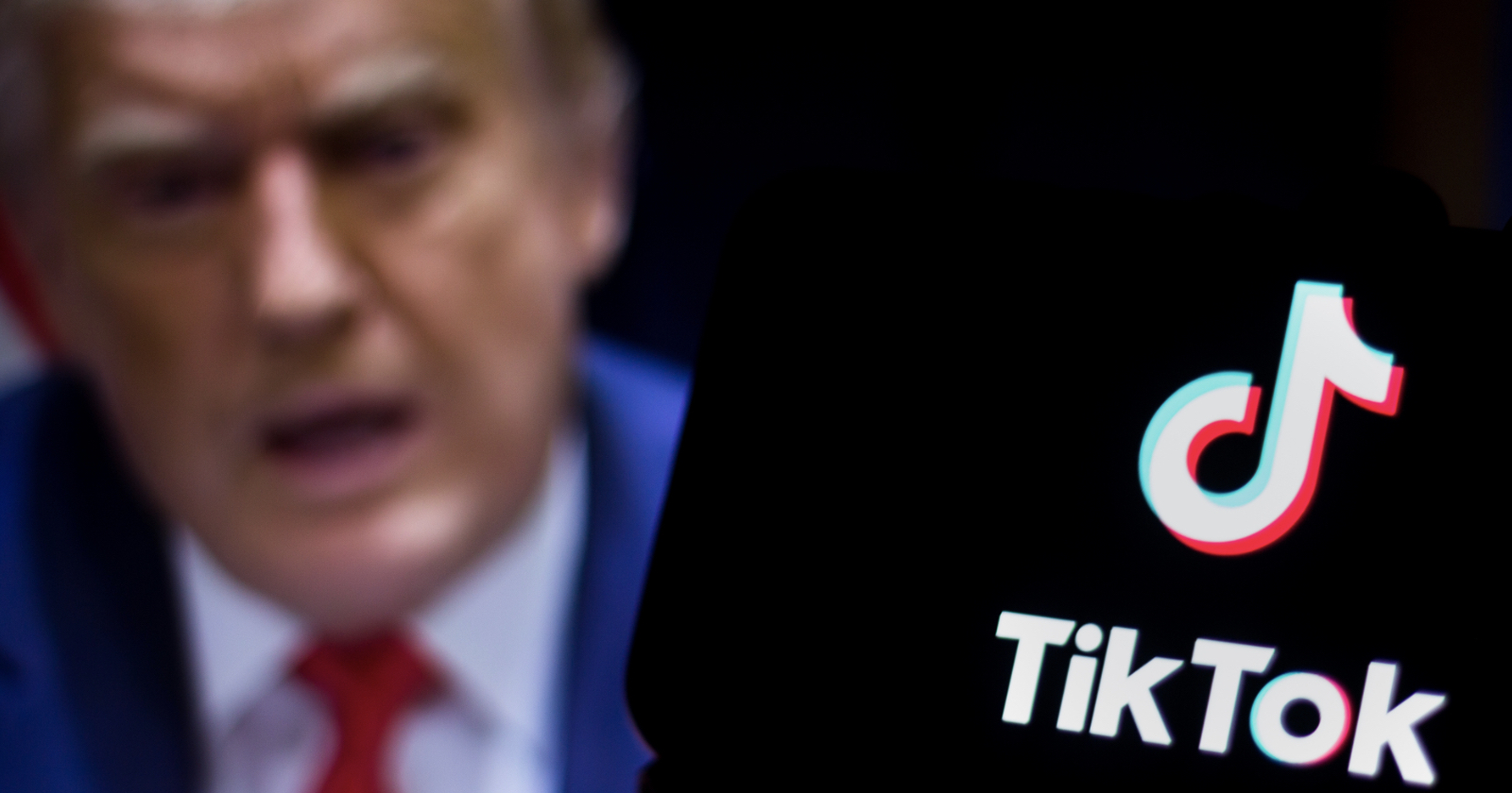 Blurred figure in a suit appears in the background, while the TikTok logo is prominently displayed in focus in the foreground. The image suggests a connection or contrast between the individual and the app.