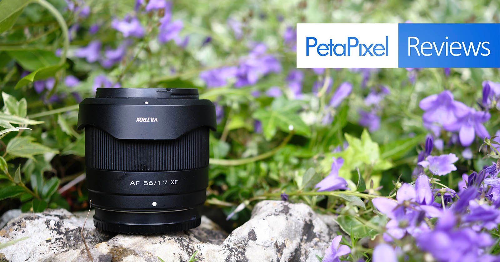 A black camera lens sits on a rock surrounded by vibrant purple flowers and green foliage. In the top right corner of the image, the PetaPixel logo is displayed alongside the word Reviews on a blue background.