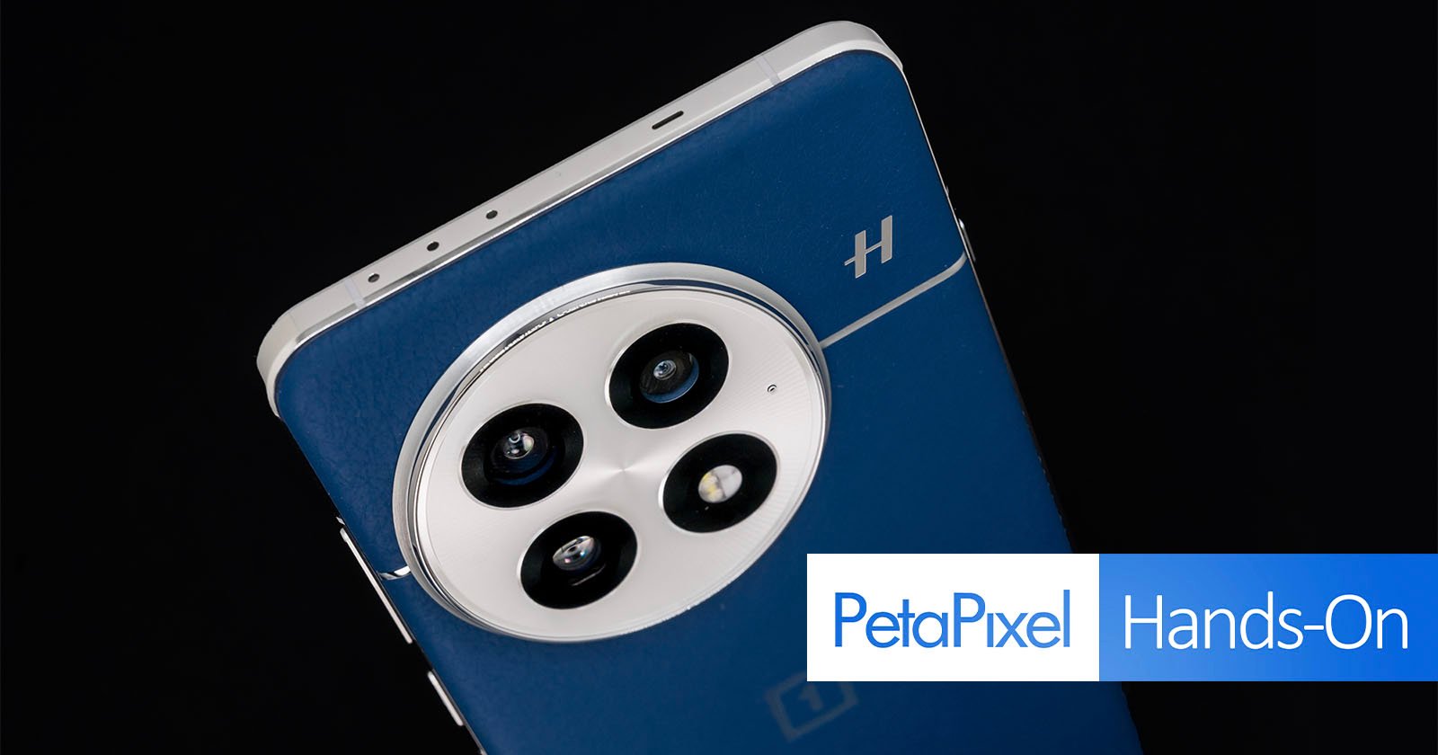 Close-up of a smartphone's rear camera module with three lenses on a sleek, dark blue back. The phone has a metallic frame and a small logo. Text overlay says PetaPixel Hands-On.