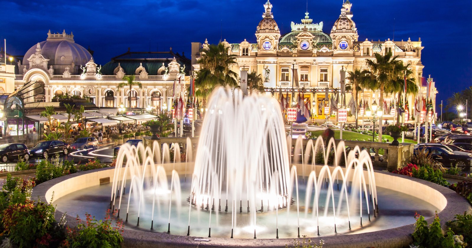 A large, illuminated fountain in front of the ornate Monte Carlo Casino at night, with palm trees, colorful flowers, and city lights in the background.