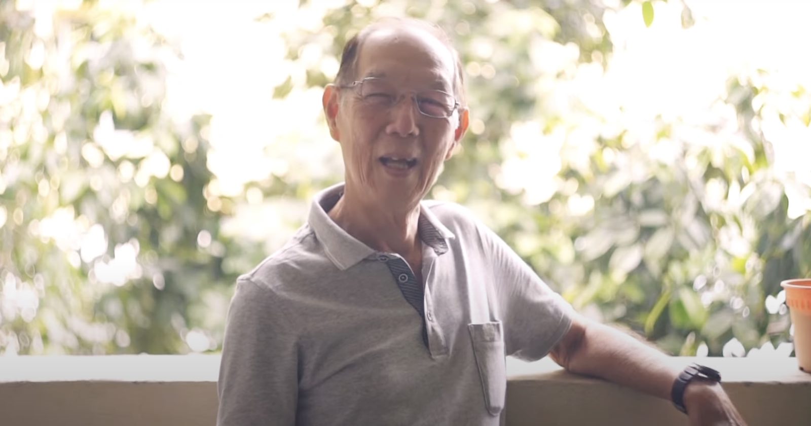 An elderly man with glasses and a grey polo shirt sits outdoors, smiling, with greenery and sunlight in the blurred background. An orange cup is on the right side of the image.