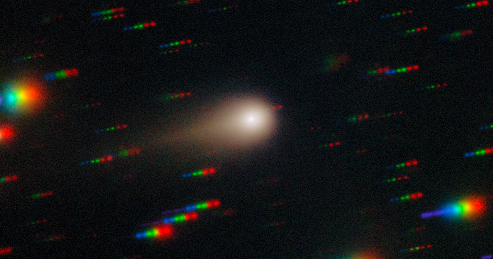 A comet with a bright core and faint tail moves through space, surrounded by colorful, streaked star trails in red, green, and blue on a dark background.