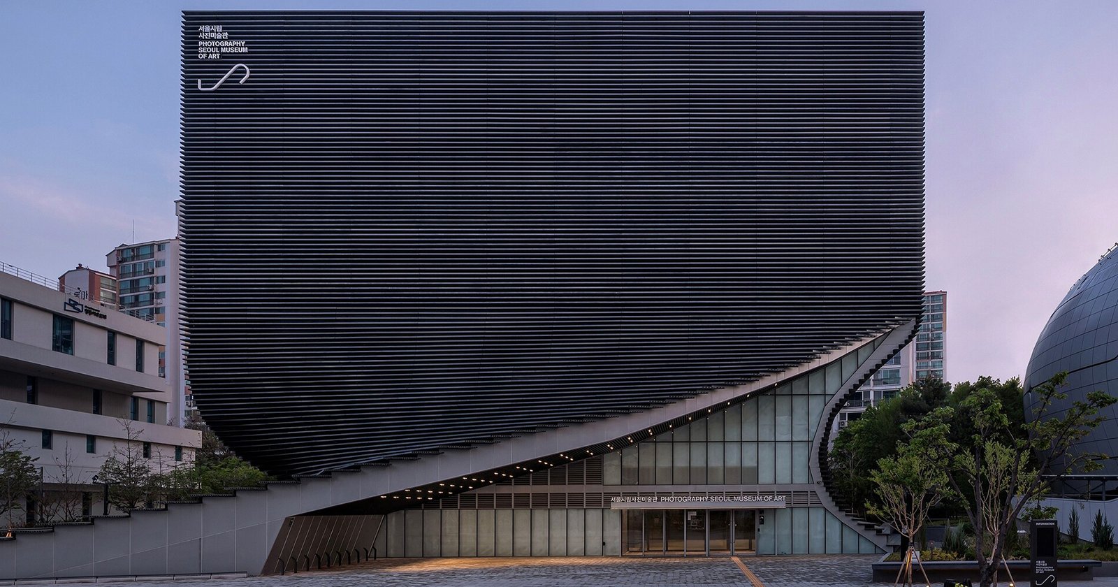 Modern building with a striking, curved facade made of horizontal black slats, partially lifted at one corner to reveal glass windows and an entrance below. Urban surroundings with trees and neighboring structures.
