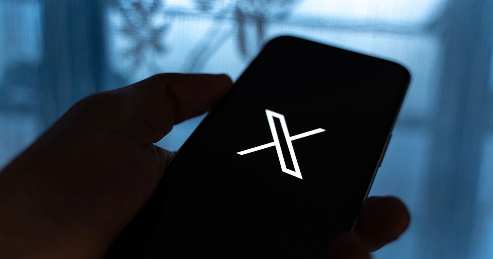 A hand holds a smartphone displaying a white X logo on a black screen, with a blurred blue background and some silhouetted shapes visible.