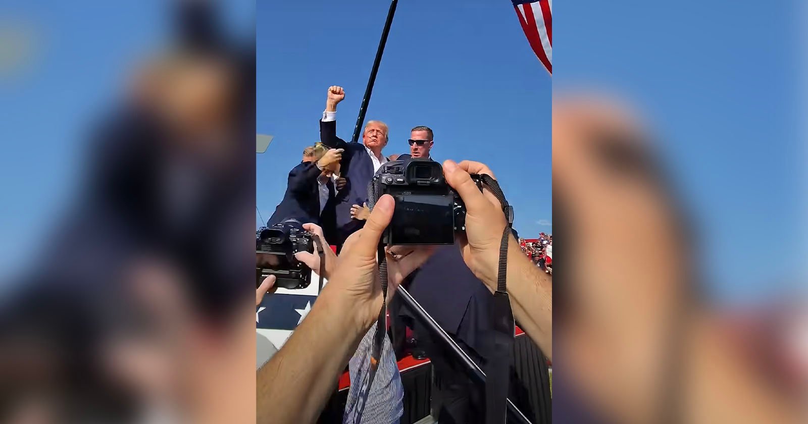 Two people are standing on a stage with one person raising a fist in the air. Several hands holding cameras are taking photos of them. It is a clear, sunny day with a blue sky in the background.