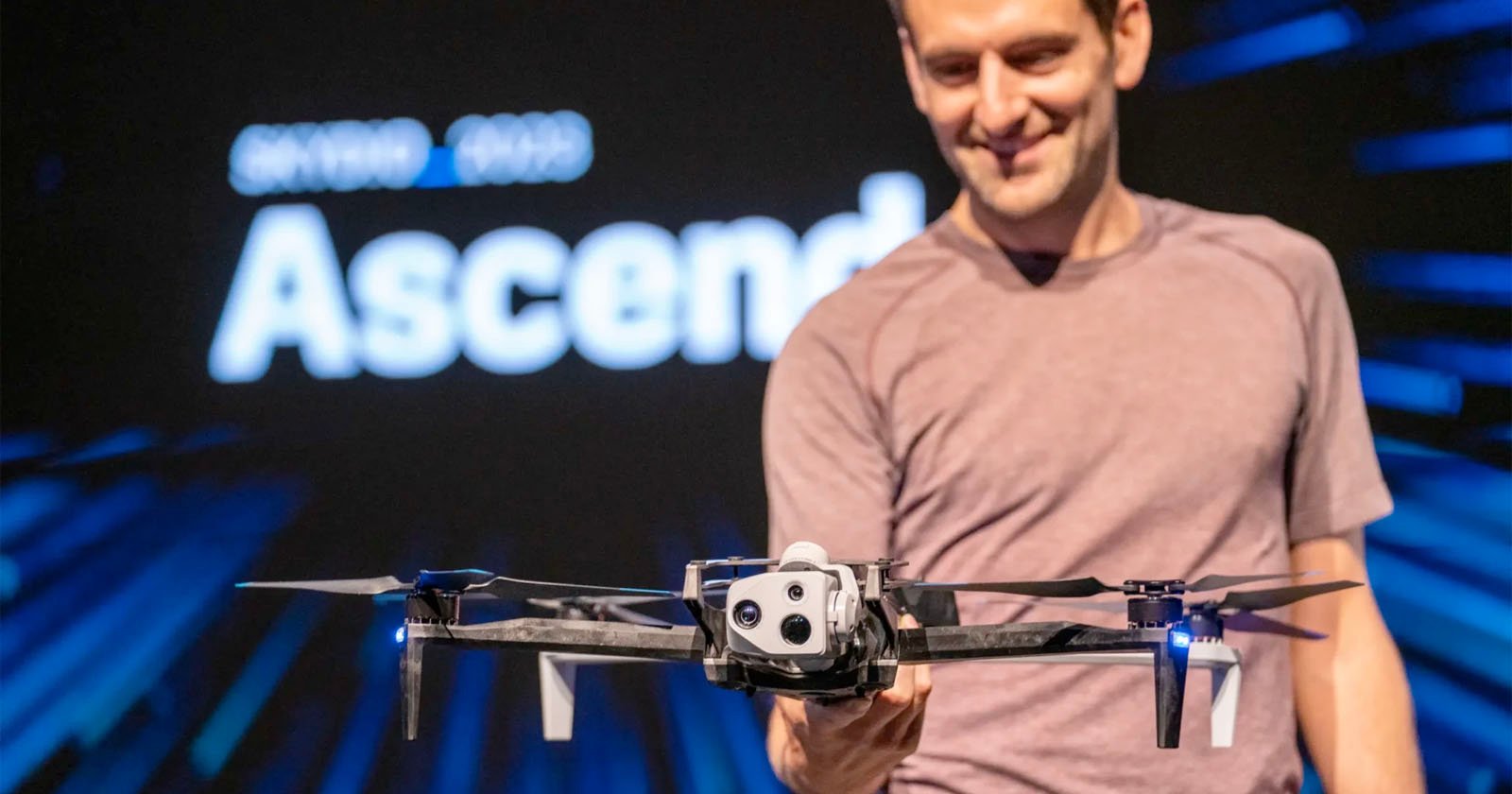 A man smiles while presenting a drone in front of a blurred background that reads Ascend. He wears a casual shirt, and the drone is featured prominently in the foreground.